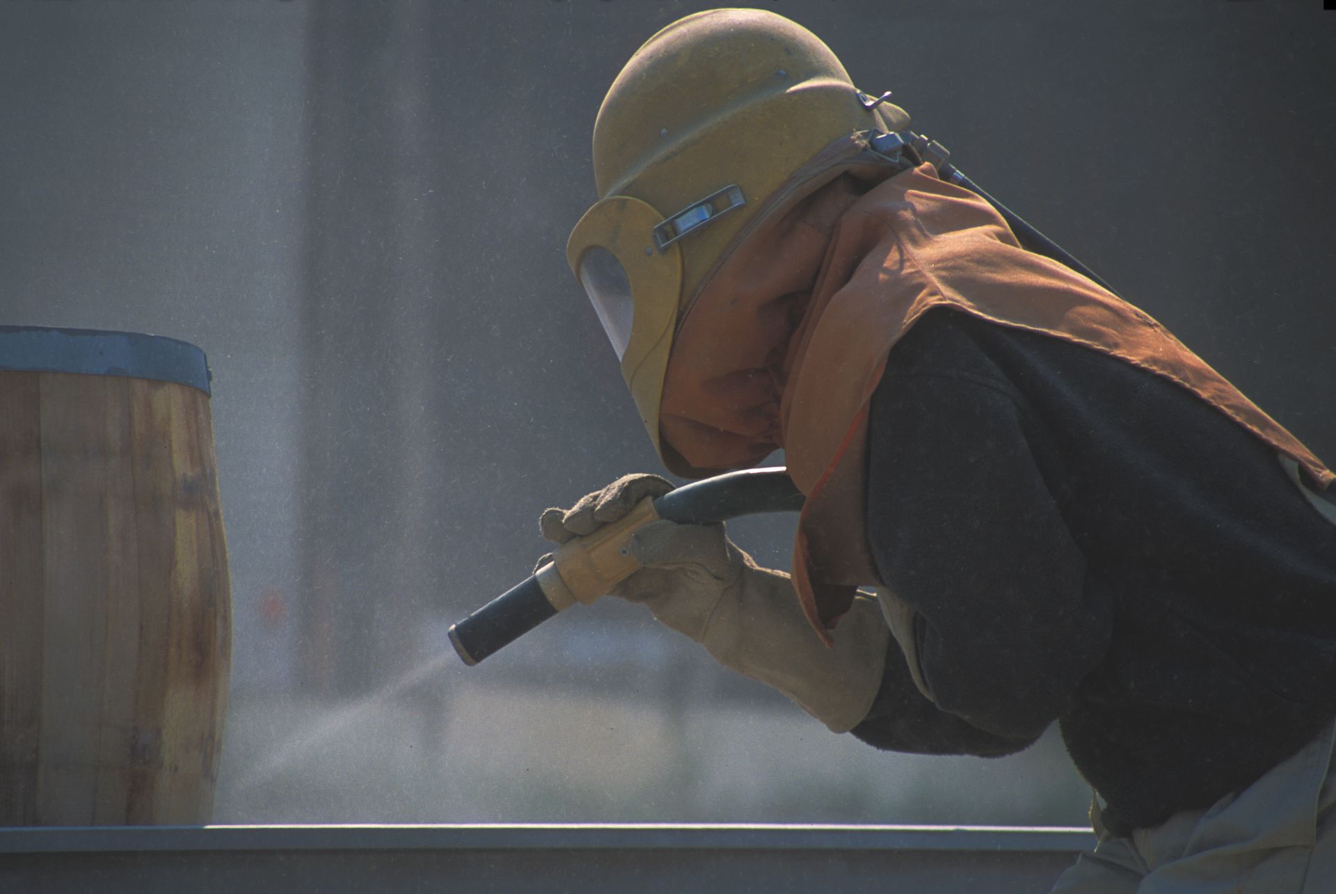 Un ouvrier portant un casque de chantier arrose un fût avec un tuyau d'arrosage dans un environnement industriel brumeux.