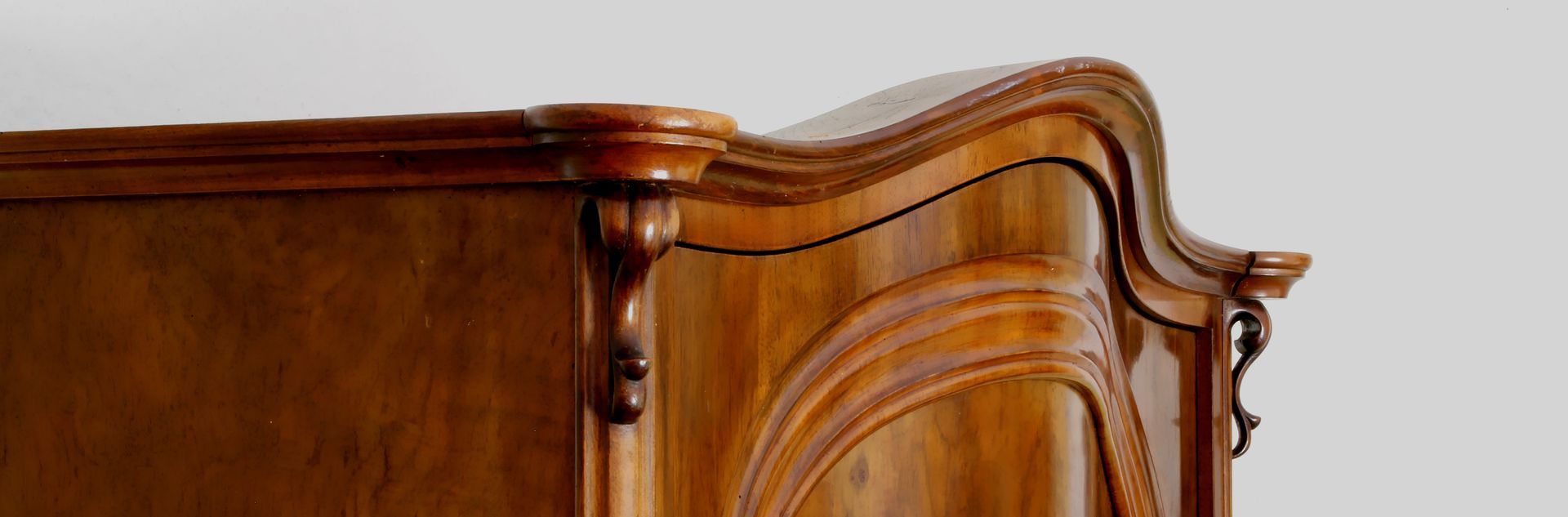 Partie supérieure d'une armoire en bois verni présentant des panneaux moulurés et une corniche aux formes courbes.