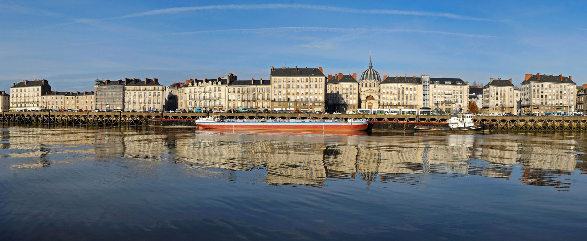Vue panoramique d'un front de rivière bordé d'immeubles, avec plusieurs bateaux amarrés et leurs reflets visibles dans l'eau.