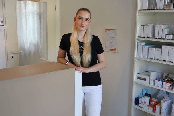 Eine Frau steht hinter einem Empfangstresen in einem hellen Büro, hinter ihr sind Regale mit Akten und Büromaterial zu sehen.
