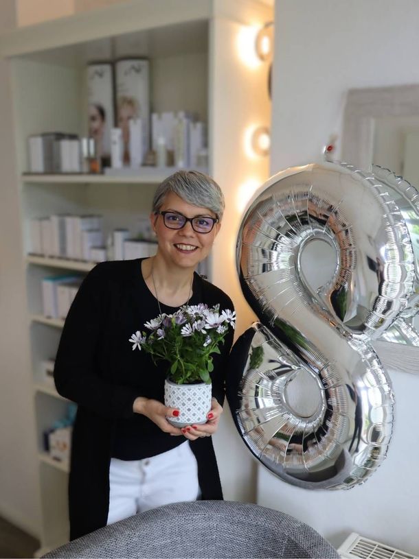 Eine Frau hält in einem hellen Raum eine kleine Topfblume neben einem silbernen, achtförmigen Ballon.