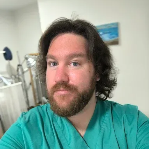 Persona con uniforme quirúrgico color turquesa tomándose una selfie en interiores, con barba y cabello hasta los hombros.