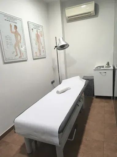 Consultorio médico con camilla de exploración, láminas anatómicas, lámpara, lavabo y aire acondicionado de pared.