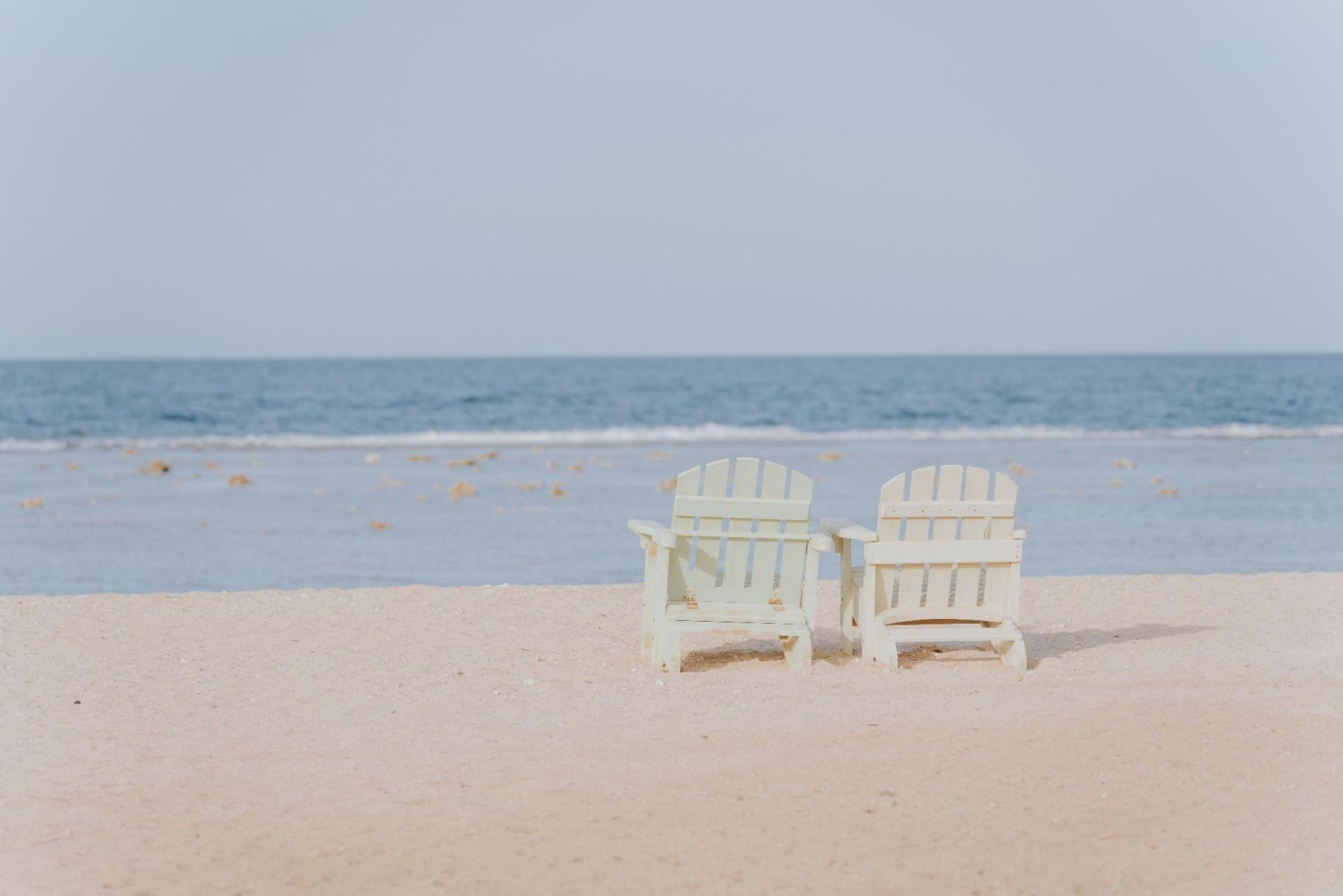 Two light-colored Adirondack chairs on a sandy beach, facing the ocean under a pale blue sky.
