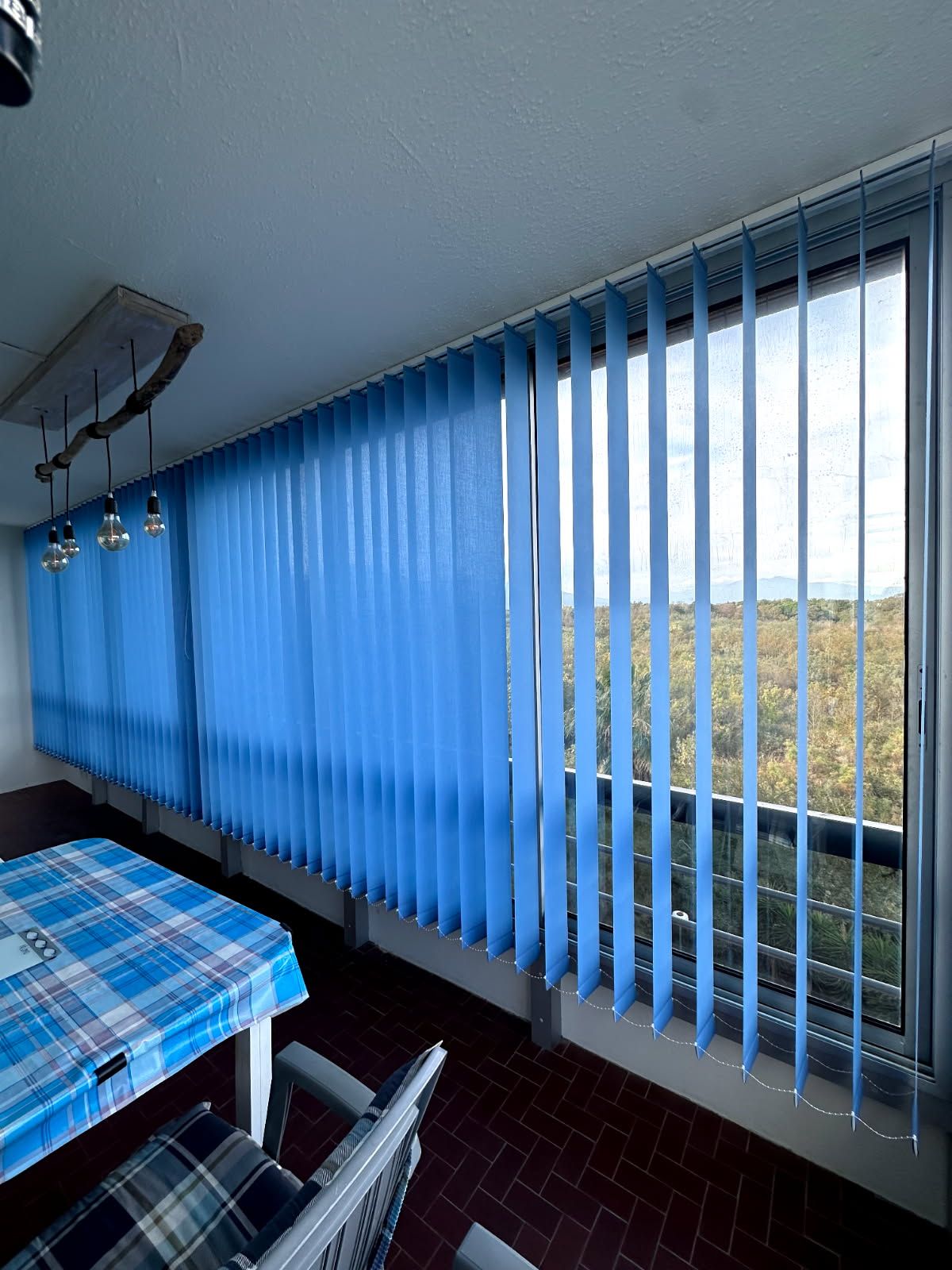 Stores verticaux bleus près d'une fenêtre dans une salle à manger, avec une table et des chaises donnant sur une vue extérieure lumineuse.