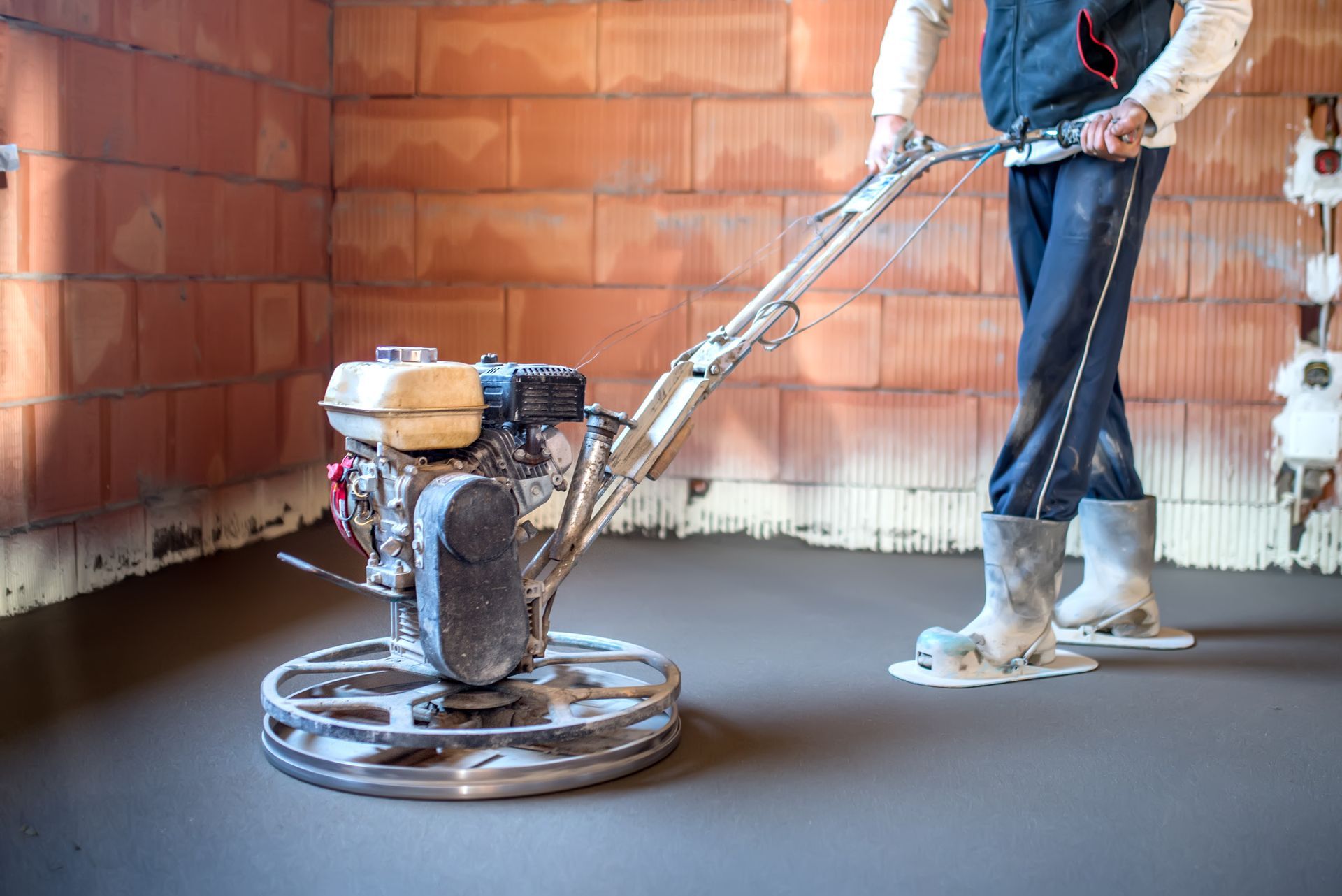 Personne utilisant une truelle mécanique pour lisser un sol en béton dans une pièce aux murs en briques.