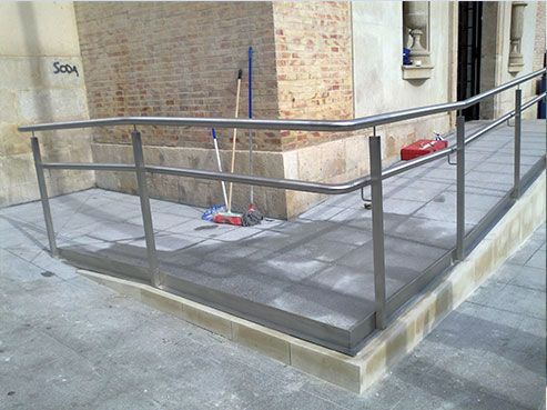 Rampa con barandillas metálicas para accesibilidad, junto a edificio de ladrillo.