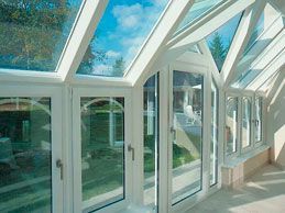 Terraza soleada y luminosa con techo de cristal y marcos blancos, con vista a un patio verde y un cielo azul.