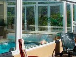 Vista de la piscina a través de grandes ventanales, con árboles verdes y la piscina visibles. Dos sillas en primer plano.