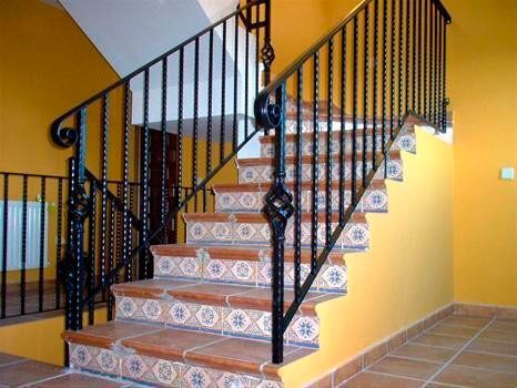 Escalera con barandilla de hierro forjado, escalones de azulejos decorativos y paredes amarillas.
