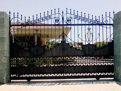 Puerta adornada de metal negro con pinchos decorativos y volutas, frente a una casa.