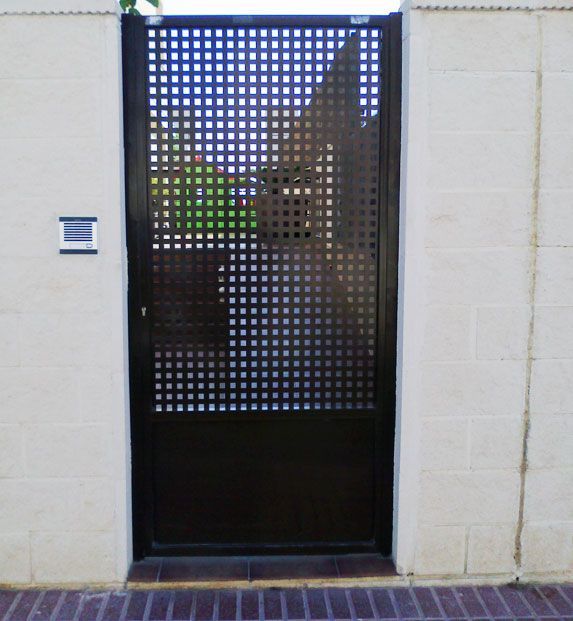 Puerta de metal negro con patrón de cuadrícula incrustada en una pared de ladrillo beige.