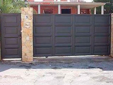 Puerta y portón de metal gris oscuro, pilares de piedra beige, frente a una casa.