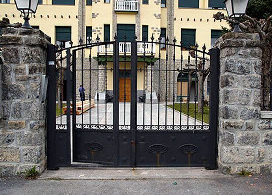Puertas de hierro negro colocadas en columnas de piedra, que conducen a un edificio con fachada amarilla.