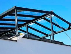 Toldo de cristal con marco negro sobre un edificio blanco, contra un cielo azul.