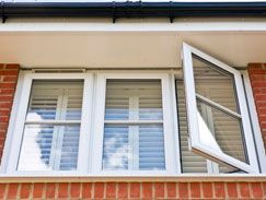 Ventana con marco blanco, con un panel abierto y persianas visibles, situada en una pared de ladrillos debajo de un edificio blanco.