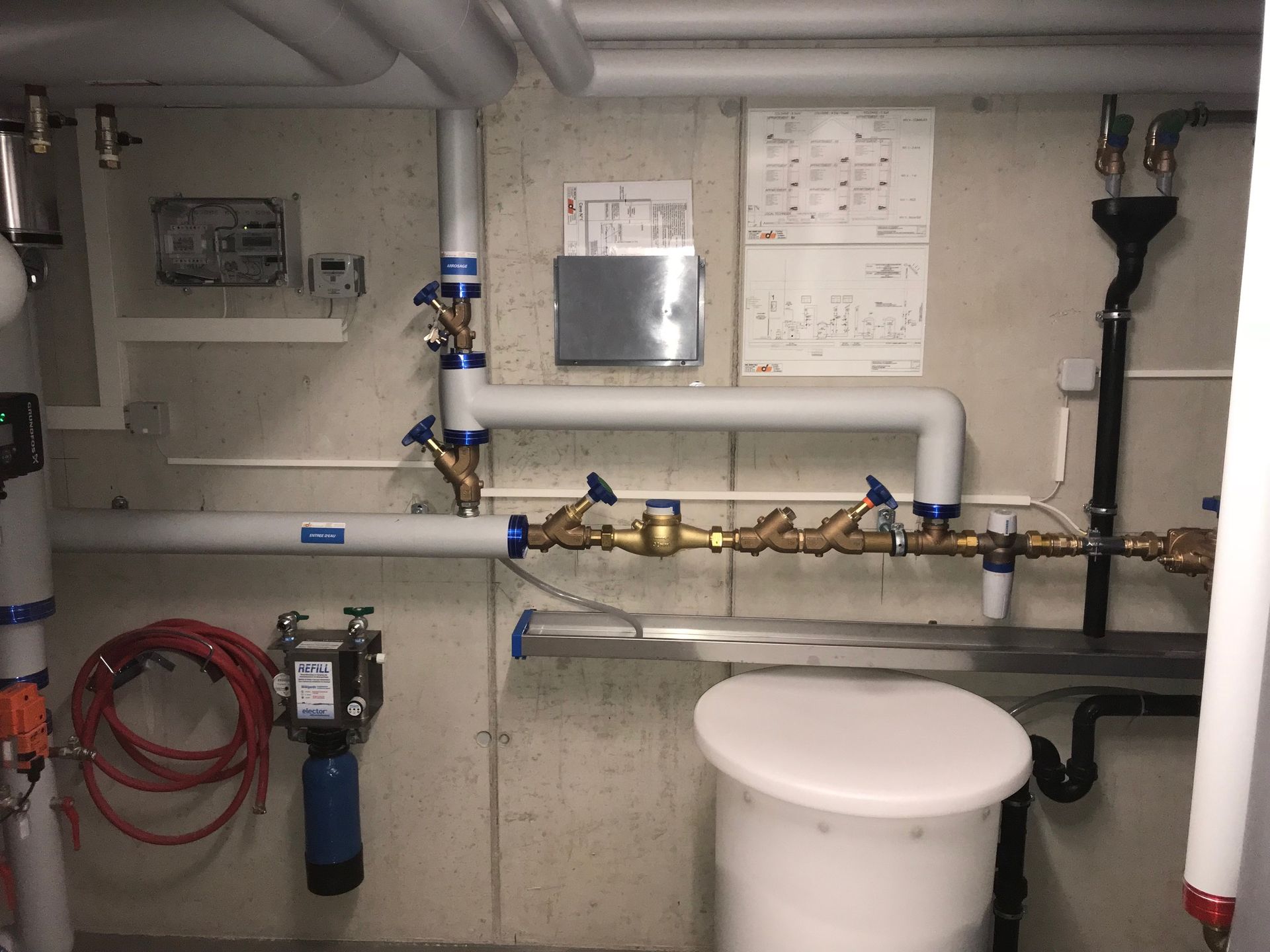 Mur de la buanderie avec tuyaux blancs isolés, vannes en laiton, compteur d'eau et petit réservoir bleu