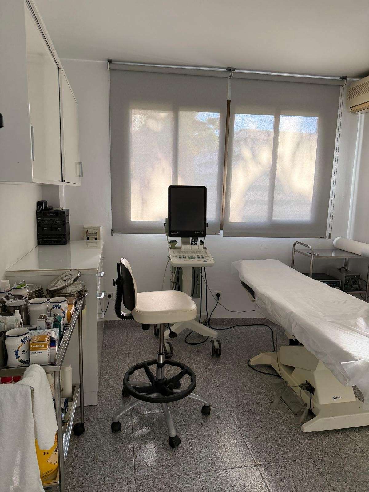 Sala de exploración médica con camilla blanca, taburete, monitor y persianas en la ventana.