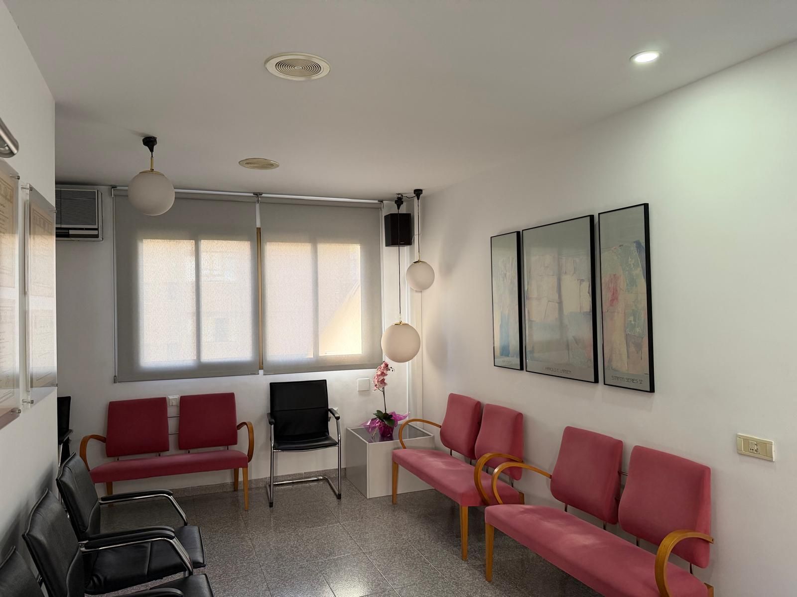 Pequeña sala de espera con sillas rojas, persianas, cuadros en las paredes y una silla de escritorio bajo luces de techo.