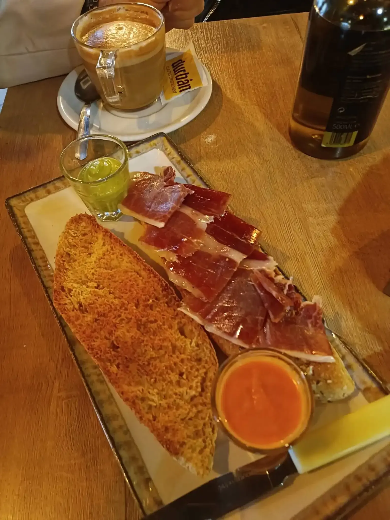 Plato de tapas con jamón en lonchas, pan, salsa y un vaso de café sobre una mesa de madera.