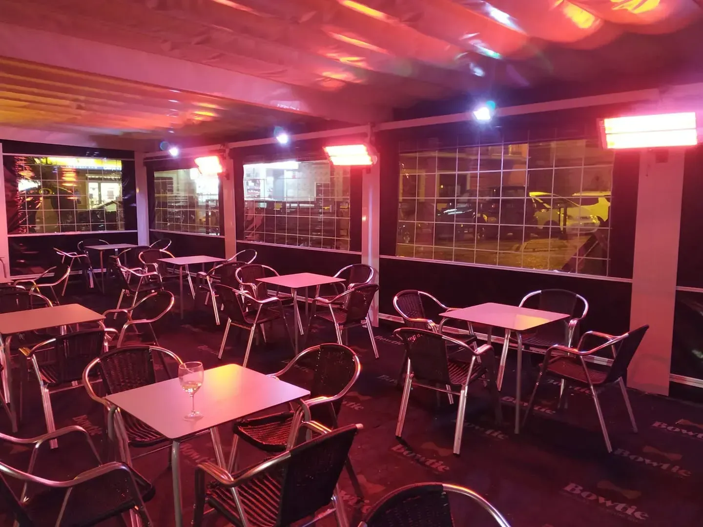 Patio de café vacío con mesas y sillas de metal bajo iluminación roja y morada por la noche.