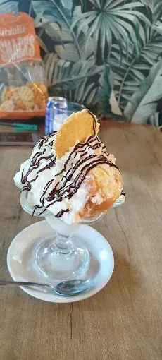Helado tipo sundae en un plato de cristal, cubierto con sirope de chocolate y una oblea, sobre una mesa de madera.