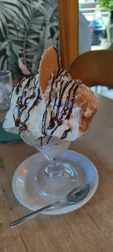Un helado tipo sundae en un plato de cristal con sirope de chocolate sobre una mesa, con una cuchara al lado.