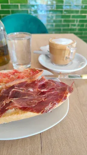Plato con pan y jamón curado sobre una mesa junto a un vaso de agua y un capuchino en una cafetería con azulejos.