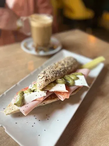 Un sándwich con semillas en un plato blanco junto a un café con leche en una mesa de madera de una cafetería.