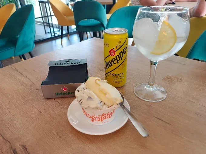 Mesa con un gin tonic, una lata de cerveza de jengibre, un pastel de postre y una caja negra en una cafetería.