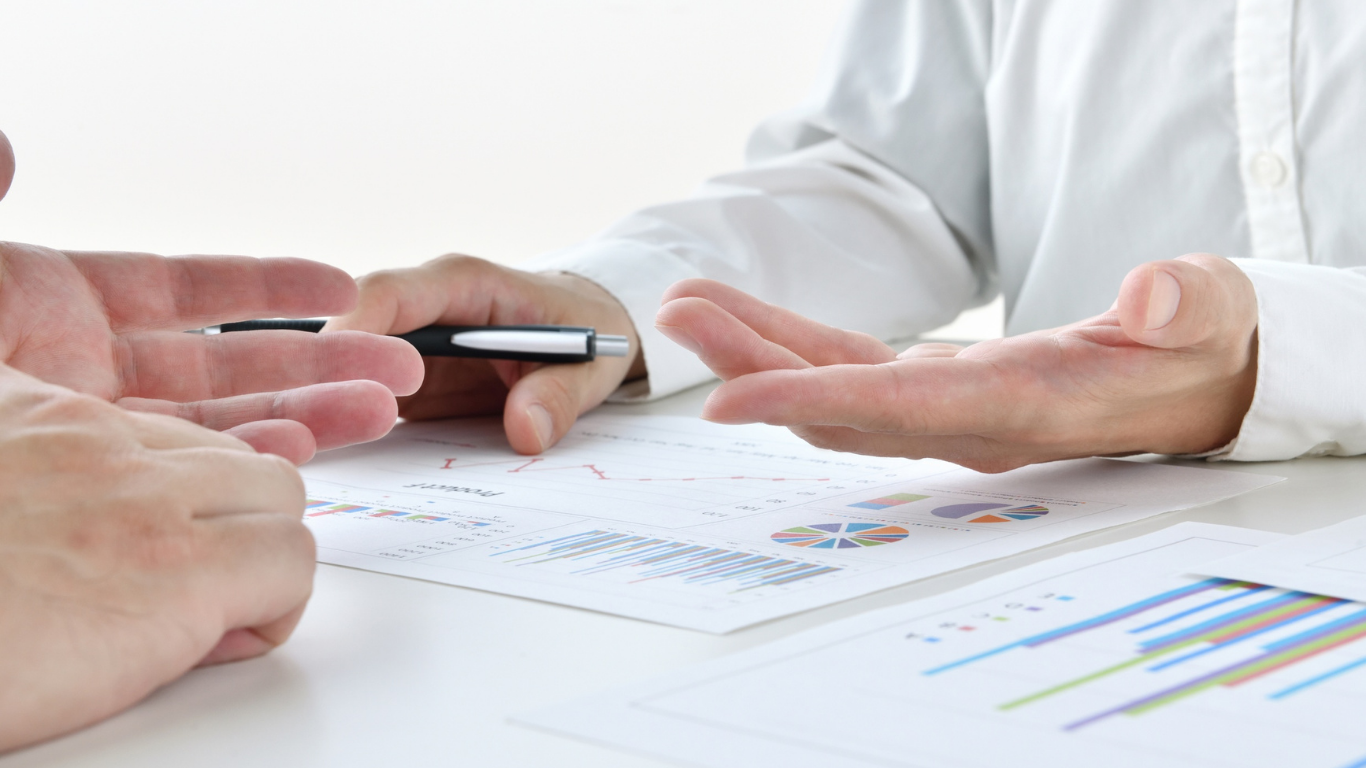 Dos personas sentadas a una mesa discutiendo gráficos empresariales, una sosteniendo un bolígrafo y la otra gesticulando con la mano.