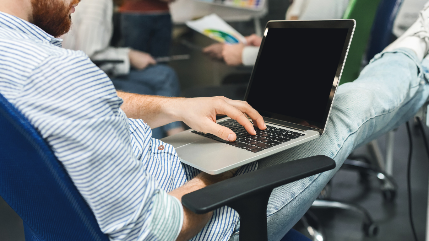 Una persona con una camisa de rayas sentada en una silla de oficina con los pies en alto, tecleando en un ordenador portátil con la pantalla en blanco.
