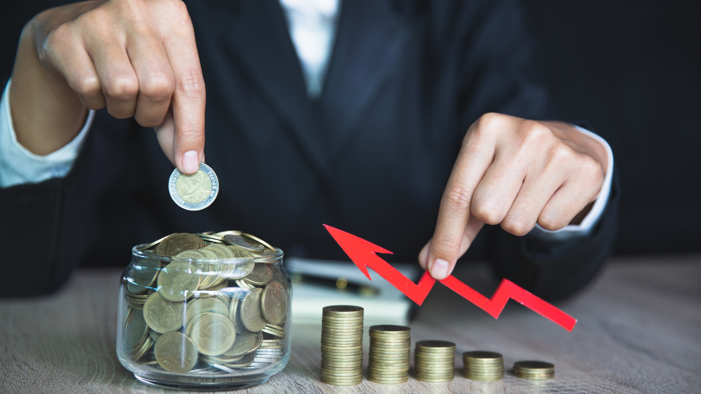 Una persona vestida con traje introduce una moneda en un frasco de vidrio junto a pilas de monedas y un gráfico de una flecha roja que apunta hacia arriba.