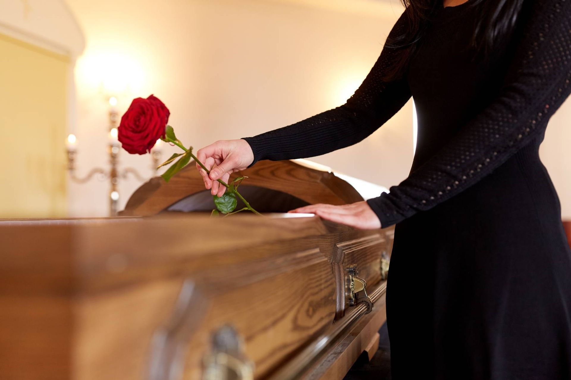 Eine Person legt eine rote Rose auf einen Holzsarg in einem sanft beleuchteten Raum.