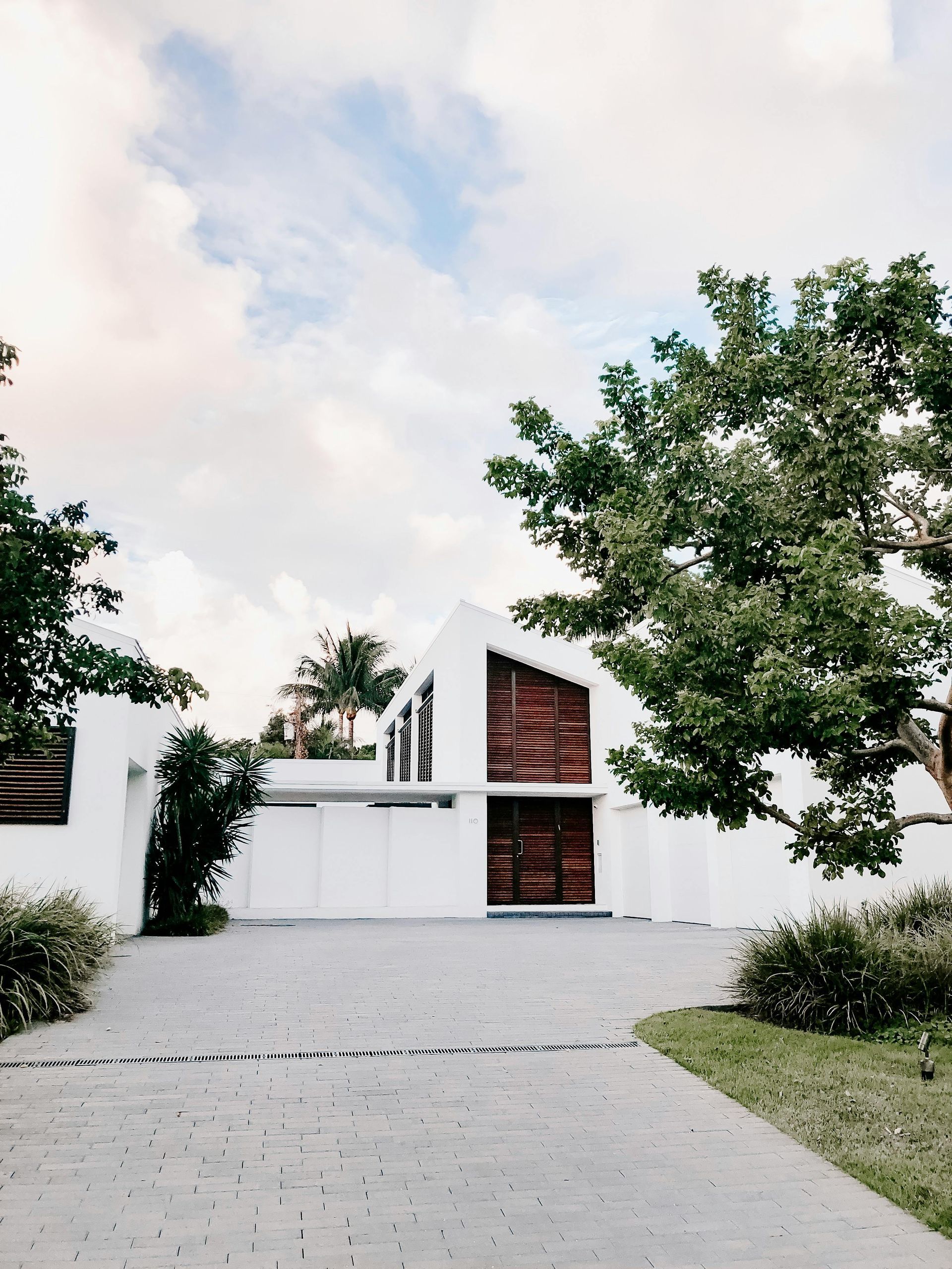 Edificio moderno blanco con puerta de madera, rodeado de árboles y un camino pavimentado bajo un cielo nublado.