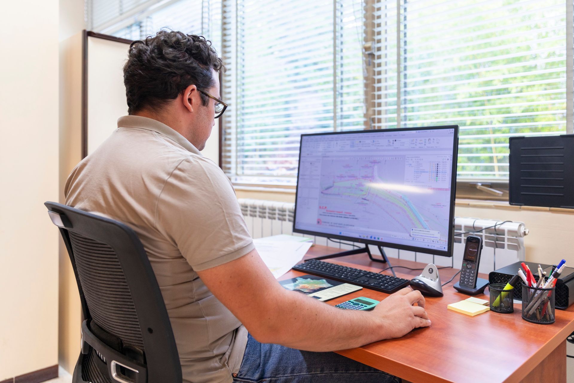 Une personne de dos devant son ordinateur en train de travailler sur un plan topographique