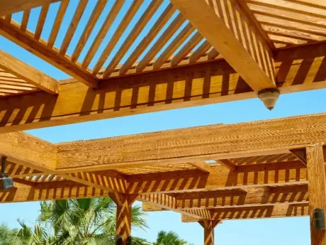 Las vigas de madera de la pérgola proyectan sombras rayadas contra un cielo azul brillante, con hojas de palmera debajo.
