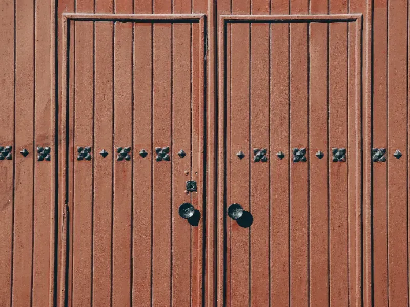 Puertas dobles cerradas de madera roja con tiradores negros y remaches decorativos de hierro.