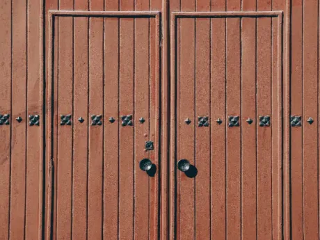Puertas dobles de madera con tiradores negros y detalles decorativos de metal sobre una pared de color marrón rojizo.