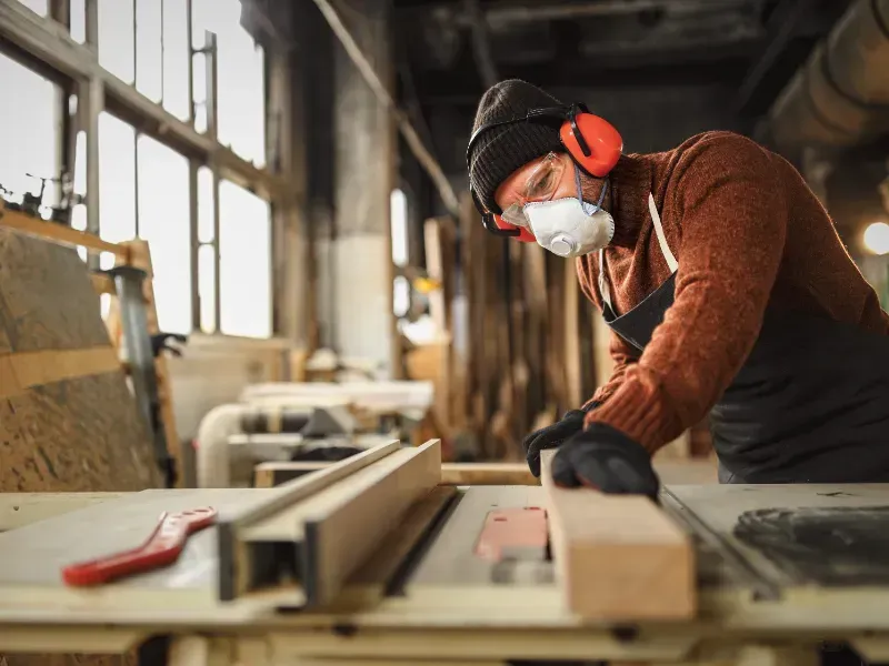 Un trabajador con equipo de seguridad mide madera en una mesa de taller, con aserrín y herramientas cerca.