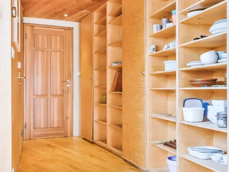 Pasillo revestido de madera con estantes empotrados para platos y cuencos, que da a una puerta de madera.