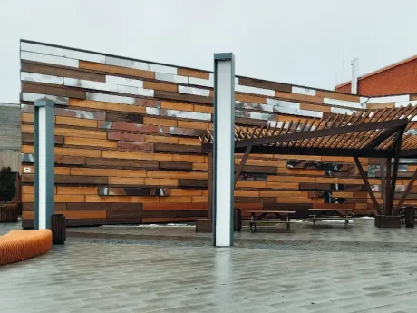 Terraza moderna en la azotea con pared de listones de madera, pérgola con asientos y pavimento mojado bajo un cielo nublado.