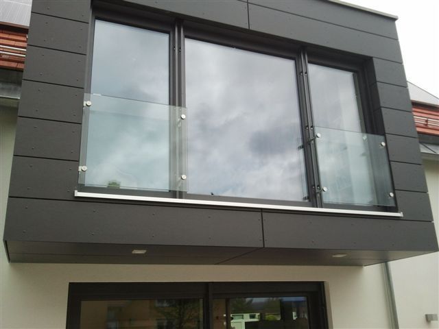 Schwarz gerahmte Fenster mit Glasgeländern an einer grauen modernen Gebäudefassade. Bewölkter Himmel spiegelt sich in den Fenstern.