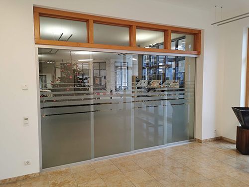 Glaseingang zum Büro mit Milchglasstreifen, Holzverkleidung und oberen Fenstern in einem gut beleuchteten Raum.