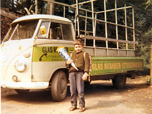 Junge im braunen Pullover mit Kegel steht vor einem VW-Truck mit Glasregal, der auf einer Einfahrt geparkt ist.