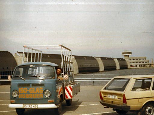 Blauer So-Glas-Kleinbus mit Glasscheiben auf einer Straße mit einem hellbraunen Auto, im Hintergrund Industriegebäude.