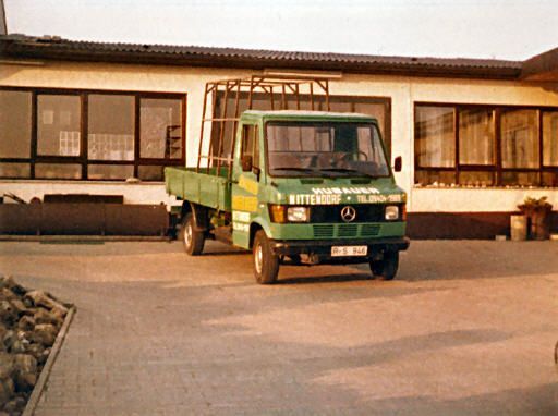 Grüner Mercedes-Pritschenwagen, geparkt vor einem Gebäude mit großen Fenstern.