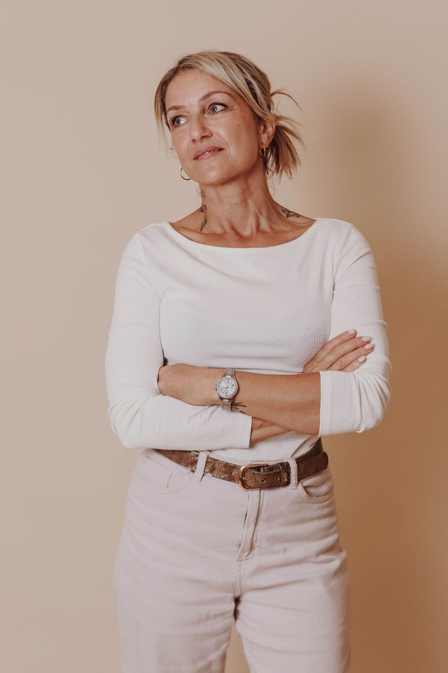 Personne en haut blanc et jean clair, debout, les bras croisés, contre un mur beige.