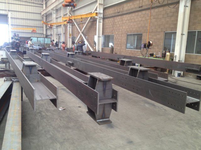 Vigas de acero en una fábrica, probablemente en construcción. Se ven trabajadores y maquinaria al fondo.