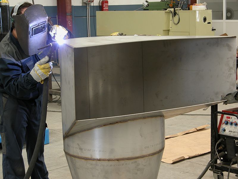 Una persona suelda una estructura metálica en un taller. Lleva máscara de soldar y guantes de protección.